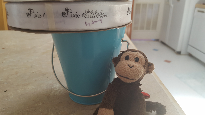 Pixie Stitches by Sway logo ribbon sitting on a small teal bucket with a plushy monkey named Playa Monkey sitting in front.