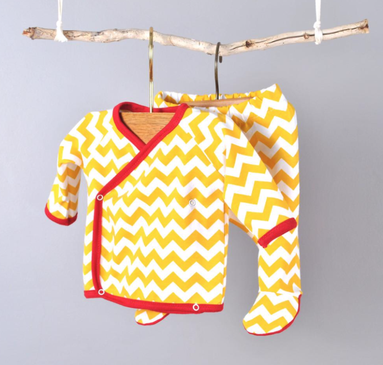 A cross-front baby shirt and a pair of fitted leggings in a zig-zag print hanging from a branch against a grey background.