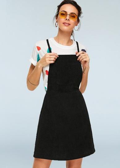 woman in a white tshirt and a black pinafore / overalls dress, front view, with hands on straps