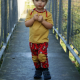 A small child in a yellow shirt and orange pants with yellow knee patches standing on a metal bridge.