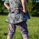 A child wearing a dark tunic length dress and purple knee length pants standing in a grassy area.