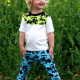 A child in a white shirt and blue and black pants standing in front of some green plants.