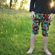 A child, shown from the mid-chest down, wearing a black, long-sleeved shirt and multi-coloured knee-length pants standing on a grassy field with the sun peeking between the child's elbow and body.