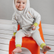A small child wearing a grey and yellow hoodie and leggings and sitting on an orange chair against a grey wall.