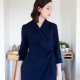 A woman in a navy blue wrap dress with three-quarter sleeves standing in front of a desk against a white wall.