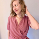 A woman in a pink, short sleeved wrap top in front of a white wall.
