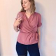 A woman in a pink, short sleeved wrap top and dark pants standing in front of a white wall.