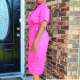 Side view of a woman in a bright pink, short sleeved wrap dress standing on a front porch.