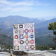 A scrappy version of the quilt is held up by a person on the edge of a rocky cliff in front of mountains and forest in the background.