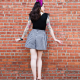 Back view of woman standing with hands to her sides in black top and wide black and white shorts in front of brick wall