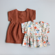 A floral baby shirt and a brown baby dress laid flat on a grey background. Both garments have a plain bodice with cut on, short sleeves and a gathered skirt.