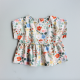 A floral baby shirt laid flat on a grey background. The shirt has a plain bodice with cut on, short sleeves and a gathered skirt.