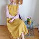 Image of a woman with short brown hair wearing a sleveeless dress with pockets in a yellow linen fabric. She is sitting on a chair and looking at the camera