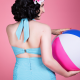 Back view of a white woman holding beachball to the side and wearing a pale blue swimsuit standing against a pink background.