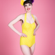 A white woman with short, dark hair wearing a yellow swimsuit standing against a pink background.