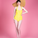A white woman with short, dark hair wearing an enormous straw hat and a yellow swimsuit standing against a pink background.