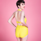 Back view of a white woman with short, dark hair wearing a yellow swimsuit standing against a pink background.