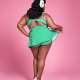 Back view of a black woman wearing a green, skirted swimsuit standing against a pink background. She's lifting the edge of the skirt on one side to show off the bottom of the swimsuit.
