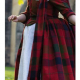 White woman dressed in reproduction 1785 clothing, including a red tartan gown, white apron, white cap, and white handkerchief around the neck. A length of the same red tartan fabric is draped over her left shoulder.