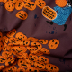Jack-O-Lantern Poplin.  A fabric printed with dark mauve background with a pile of orange jack-o-lantern pumpkins with a sign reading &amp;quot;Pumpkin Carving Contest&amp;quot;. A skeleton trick-or-treater in and oversized blue shit is holding a jack-o-lantern to the right of the pile