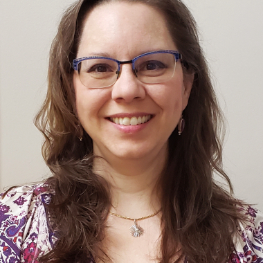 Photo of woman with long brown hair and glasses, wearing a purple shirt and smiling.