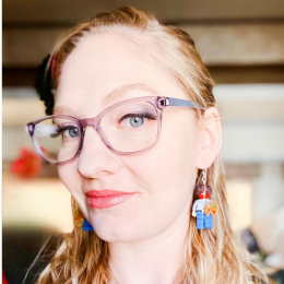 A woman's face, with blonde hair, glasses and Lego earrings.