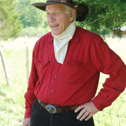 Francis Chester stands outside wearing black cowboy hat, beige bandana, red shirt and black pants.